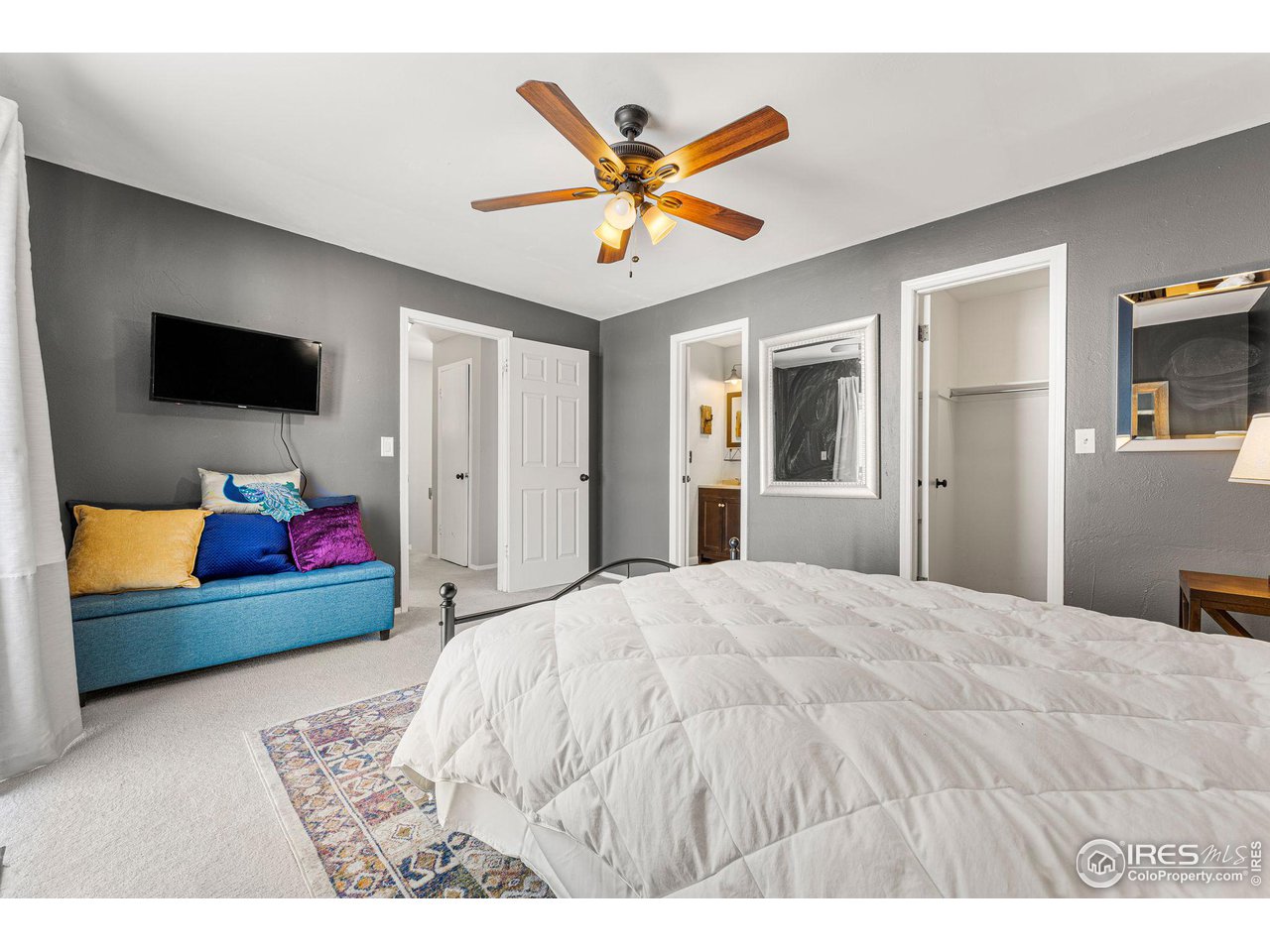 Bedroom with a white-covered bed, blue loveseat, wall-mounted TV, and wooden ceiling fan.