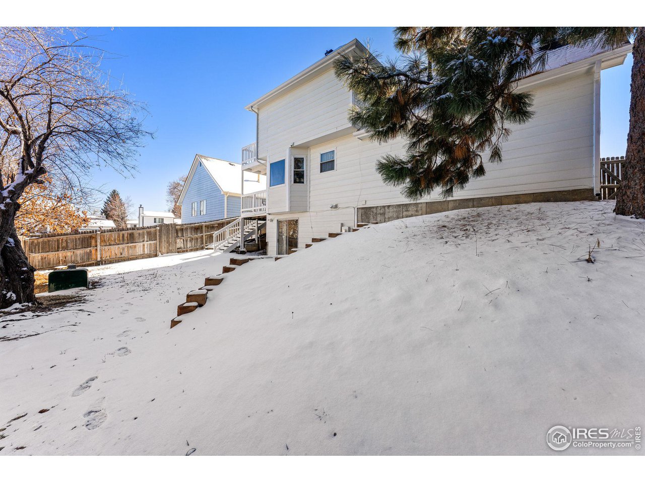 White-sided house beside a snow-covered sloping yard, wooden steps, and a fenced backyard.