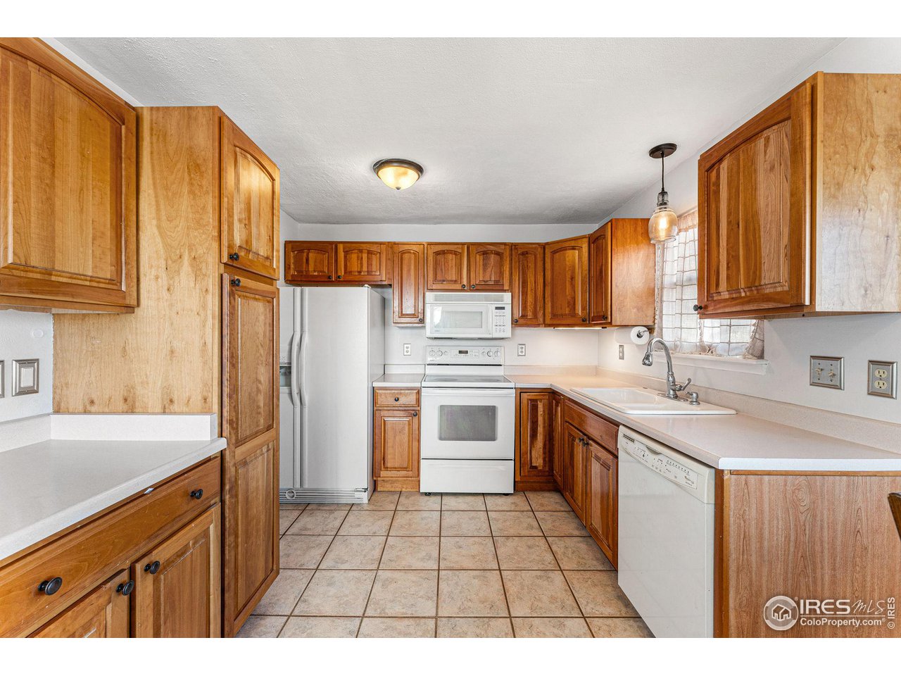 Kitchen with honey-toned cabinets, white appliances, tiled floors, and a double sink beneath a window.