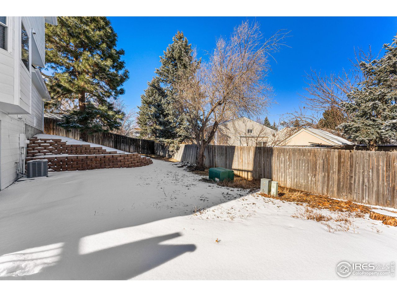 Snow-covered backyard with a tiered brick retaining wall, wooden fence, and evergreen trees.
