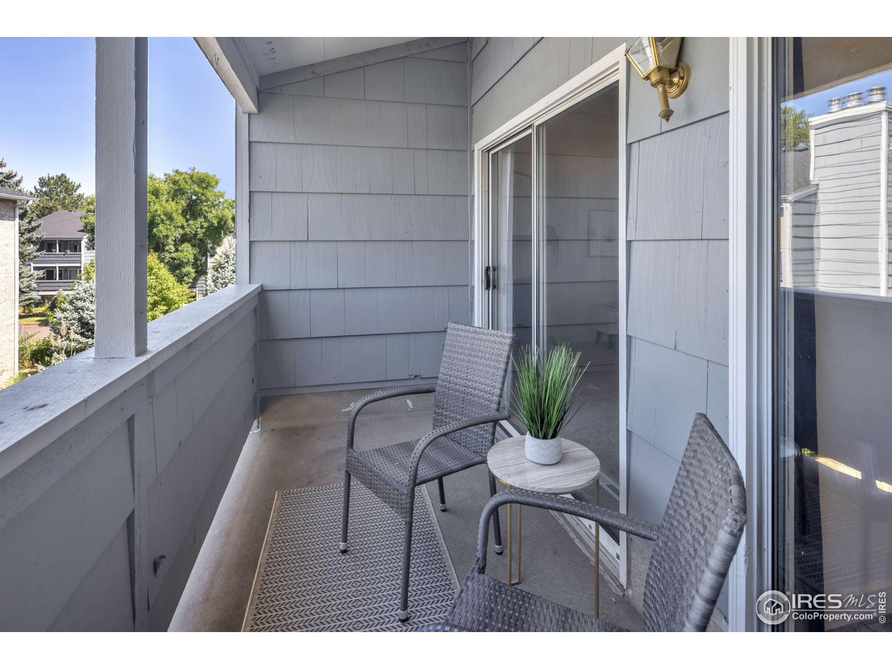 Covered balcony with woven chairs, a small table, and potted greenery beside sliding glass doors.