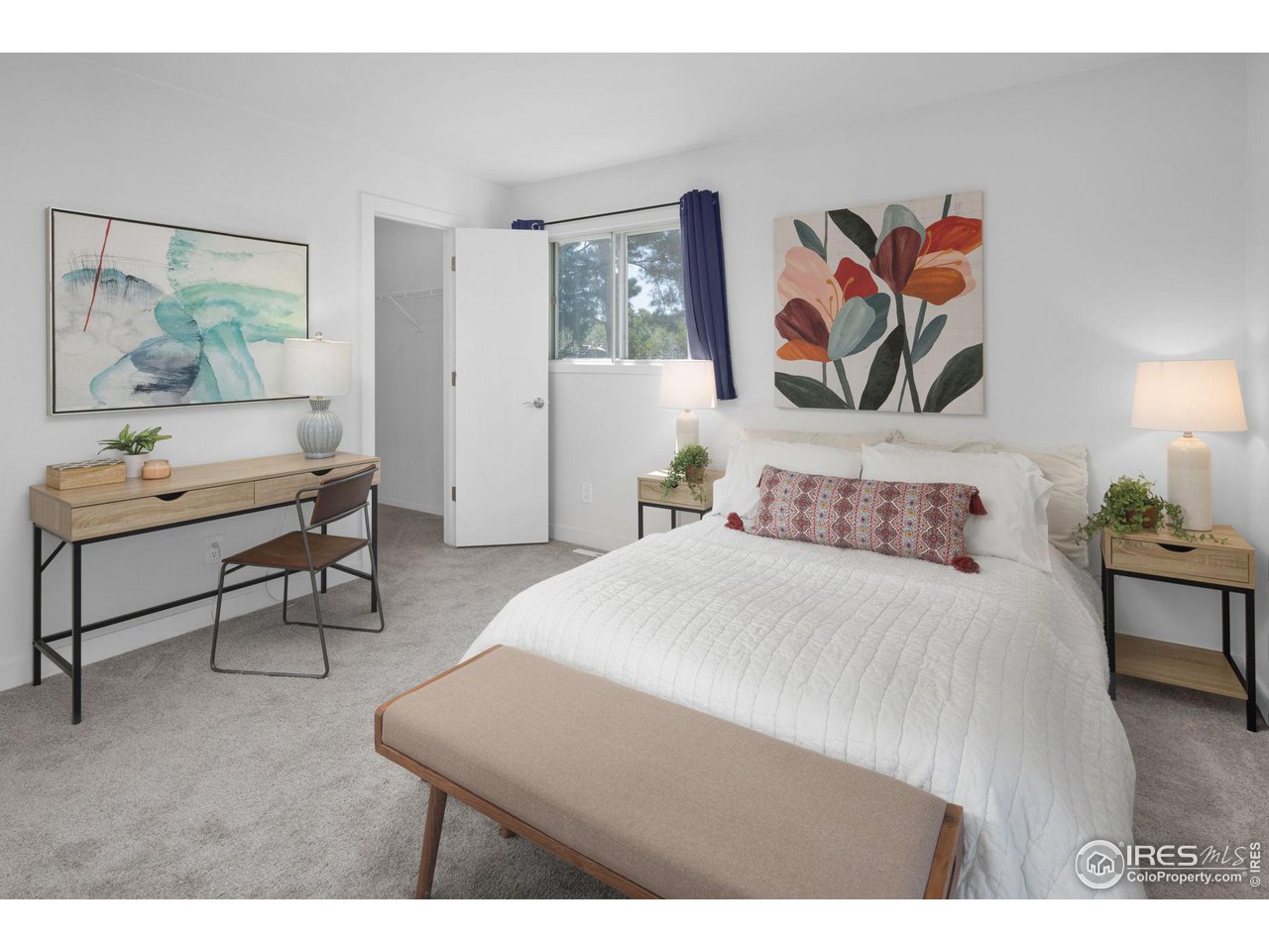 Staged bedroom with a white bed, colorful floral artwork, and a desk beside an open closet.
