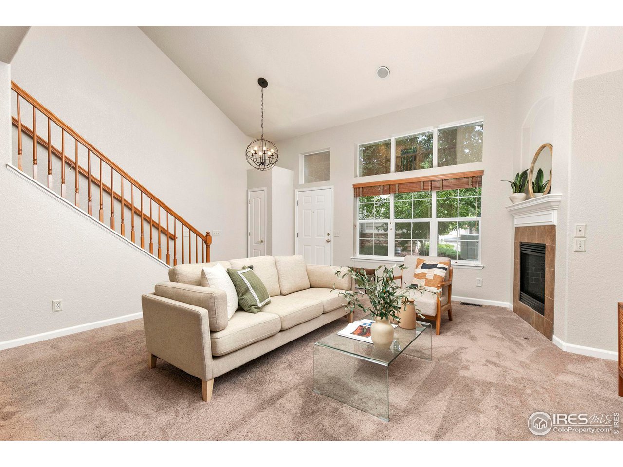 Furnished living room with vaulted ceiling, large windows, staircase, and fireplace.