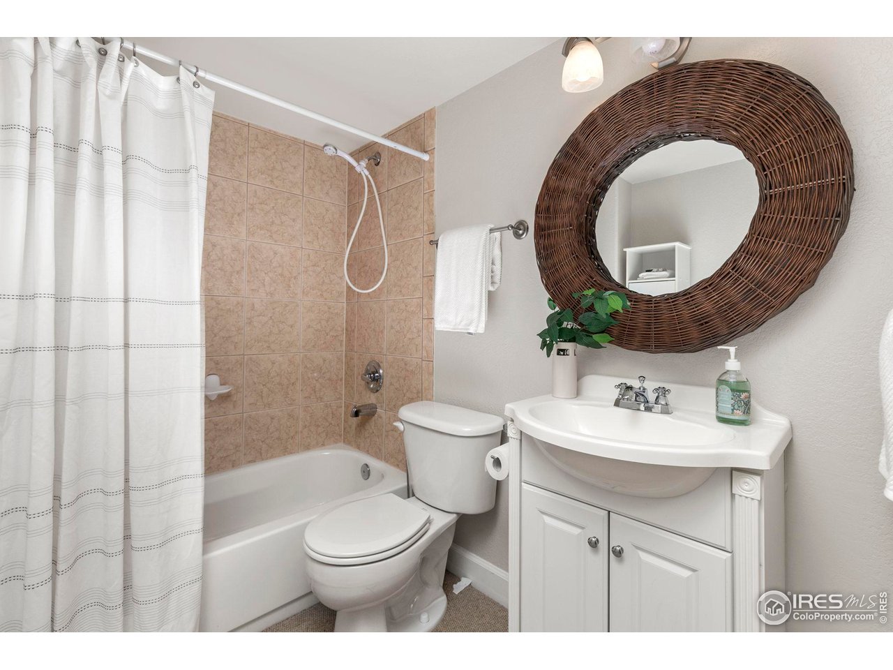Bathroom with a white vanity, large woven round mirror, and tan tiled bathtub surround.