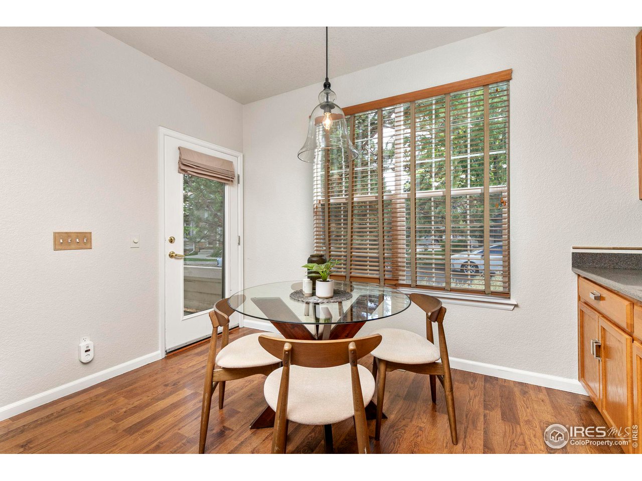 Dining nook with a round glass table, four wooden chairs, and a large window with wooden blinds.