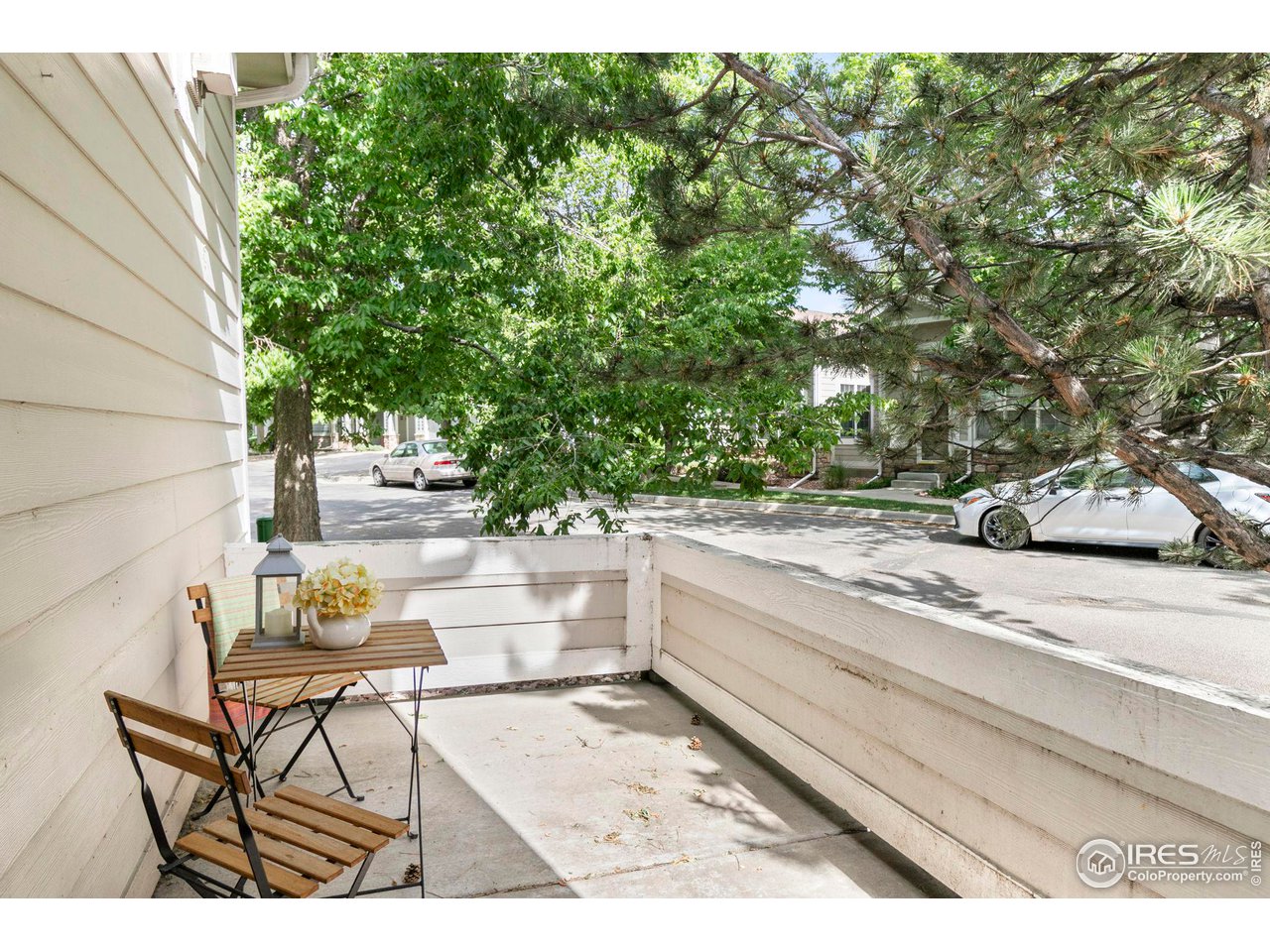 Concrete patio with folding chairs and a flower-topped table beneath leafy trees.