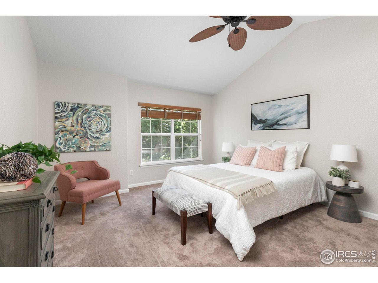 Staged bedroom with a white bed, blush pink chair and pillows, and a wood-blade ceiling fan.