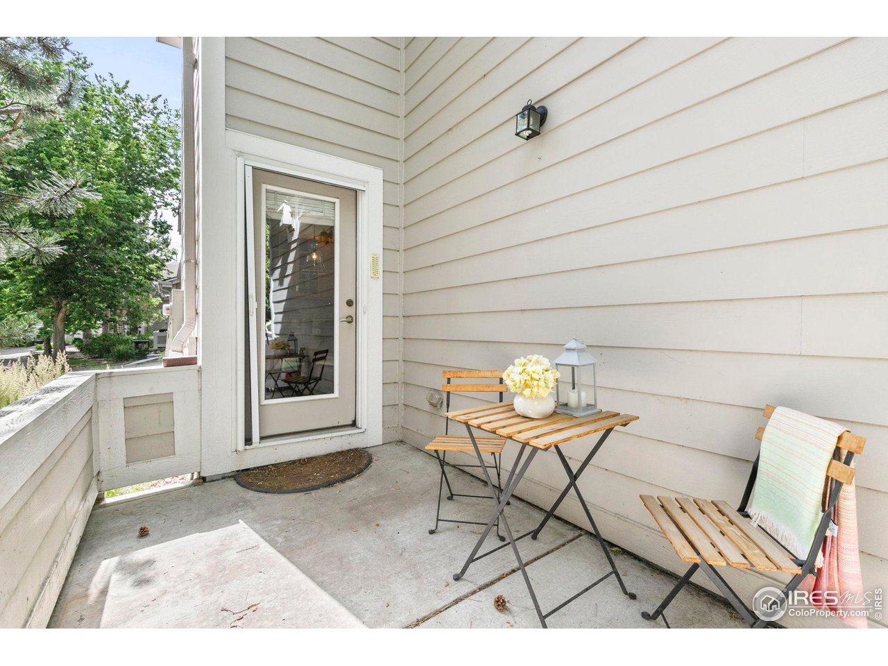Covered patio with a glass-panel door, wooden bistro set, and flowers beside beige siding.