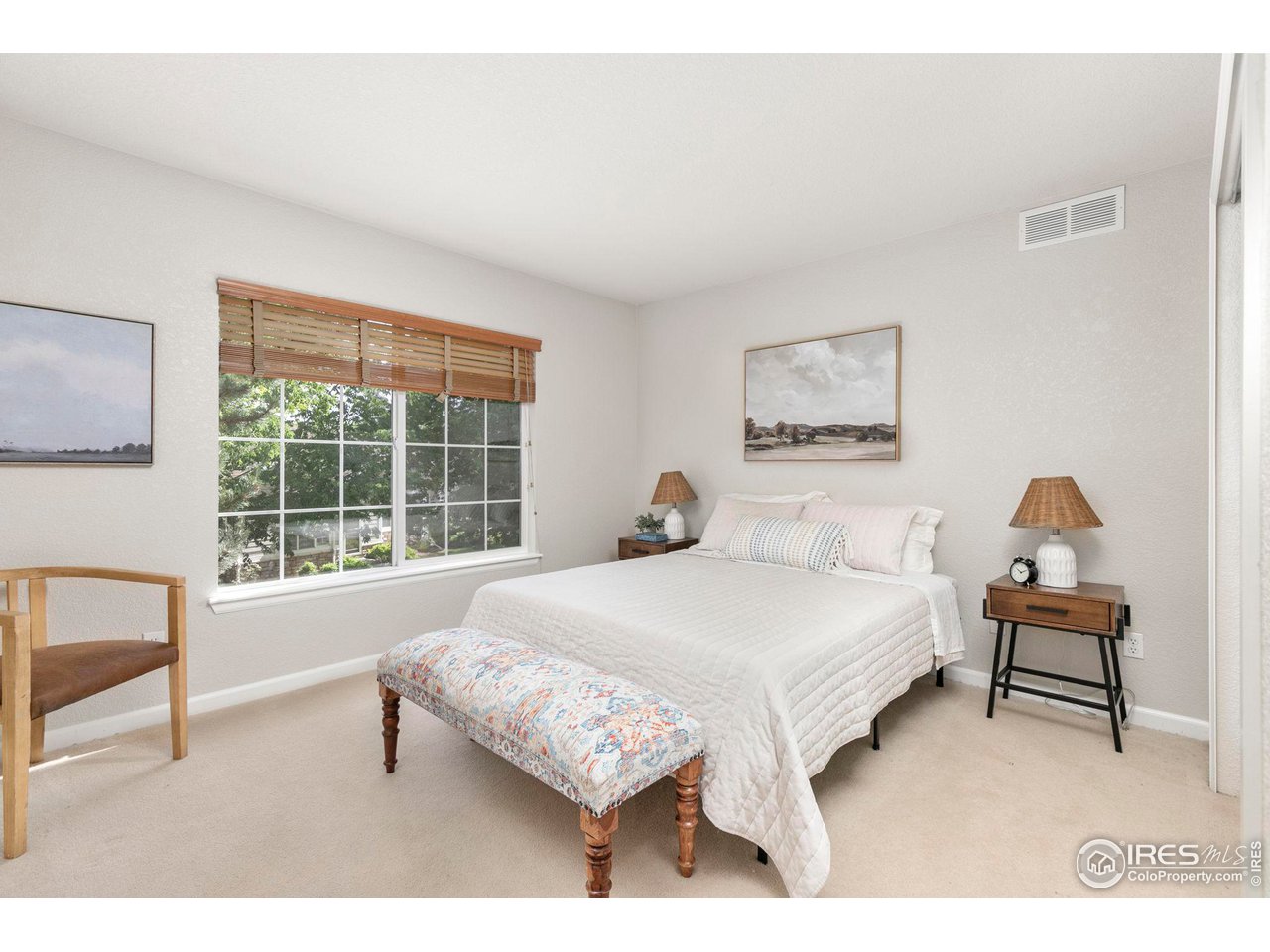 Furnished bedroom with a white bed, patterned bench, and large window with wooden blinds.