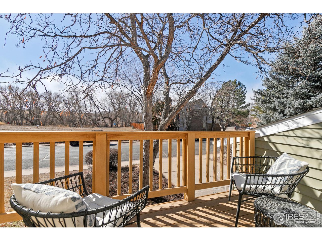 Wooden deck with woven chairs and cushions overlooking a residential street and leafless trees.