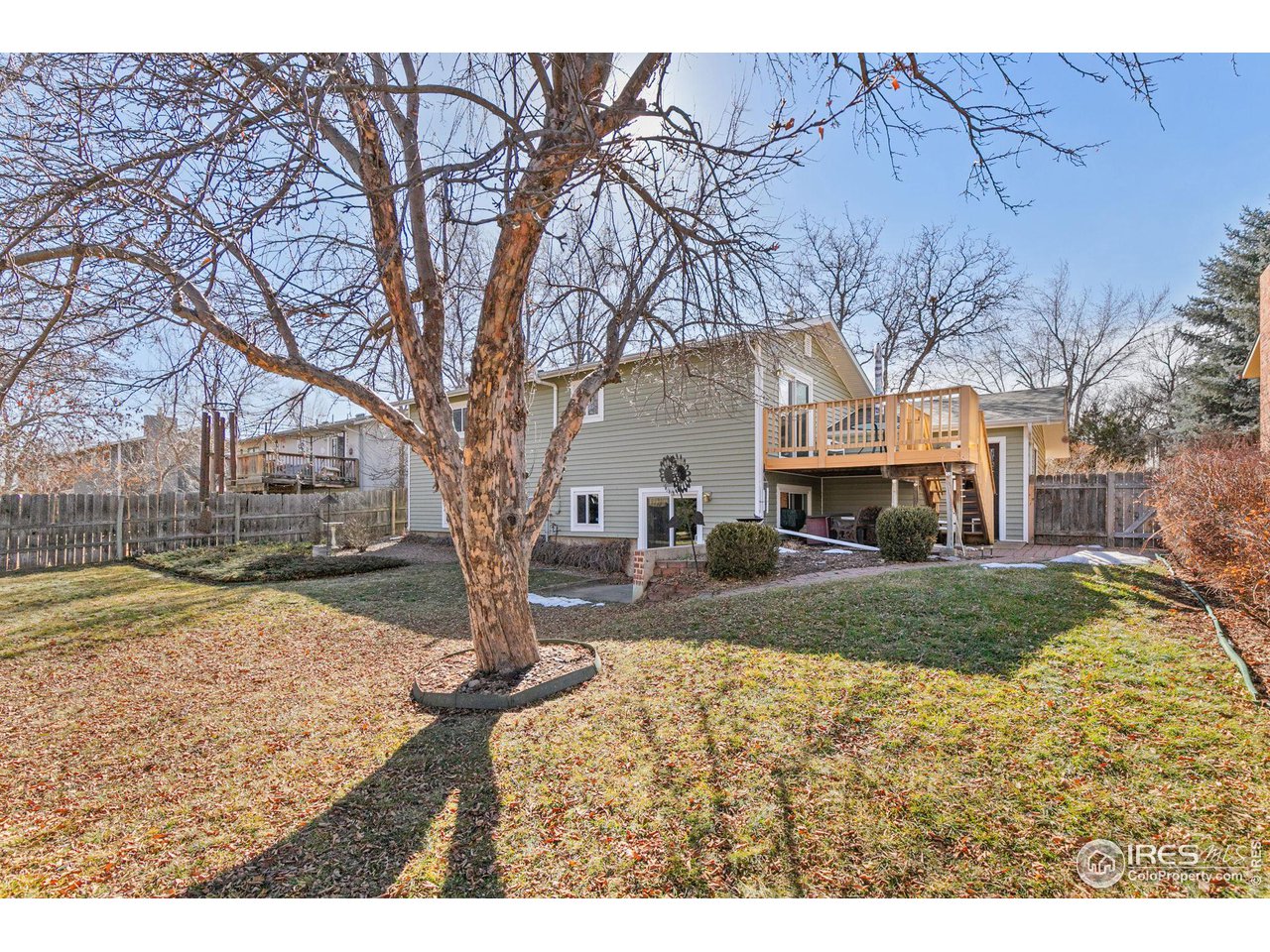Rear of a gray two-story house with a raised deck, seen across a backyard with a large leafless tree.