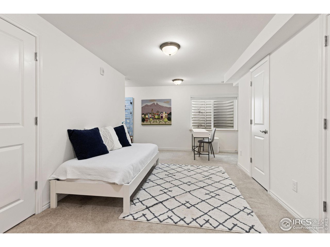 Furnished bedroom with a white daybed, geometric rug, and small desk beneath a shuttered window.