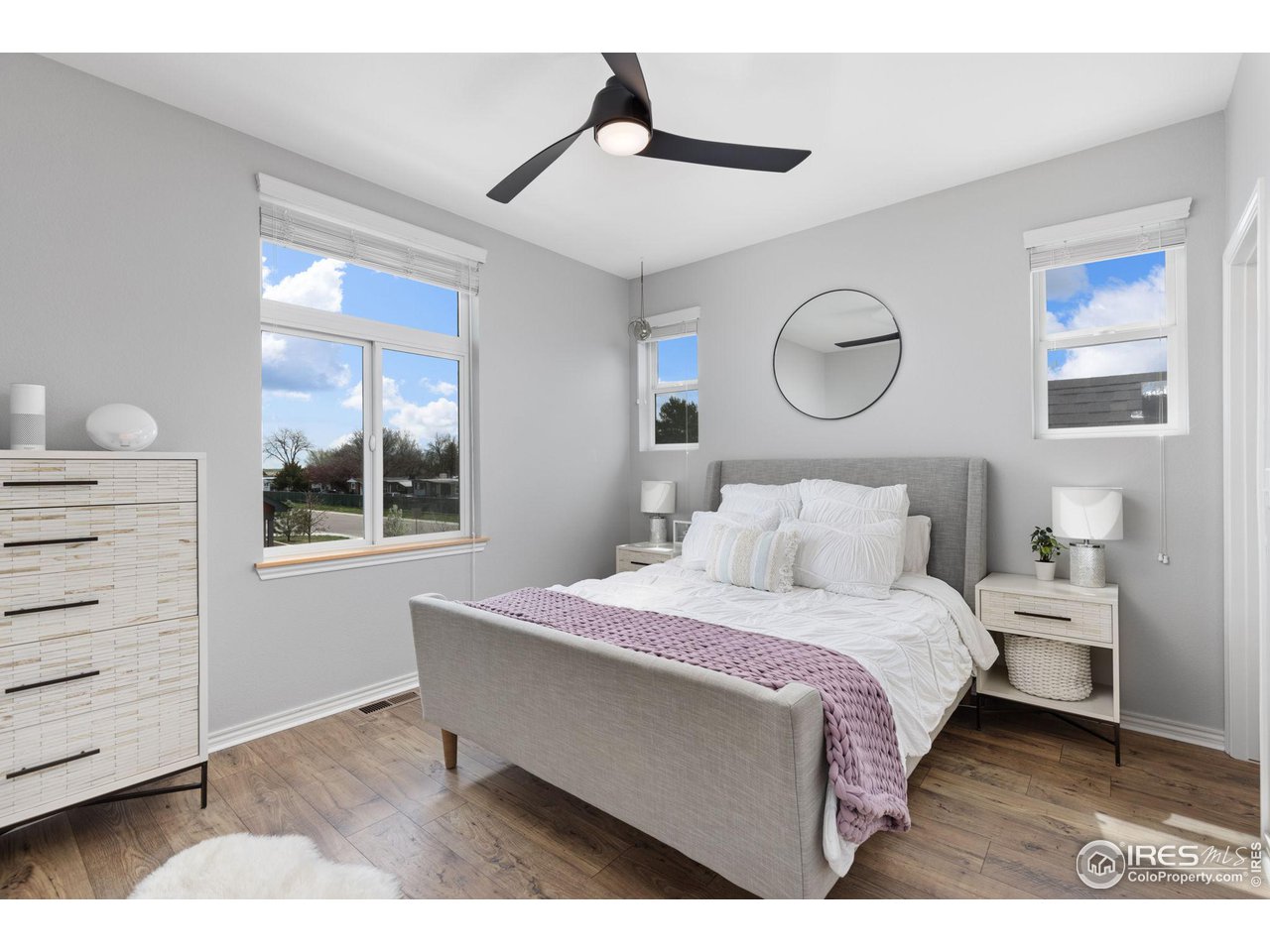 Staged bedroom with a gray upholstered bed, mauve throw, round mirror and windows on three sides.