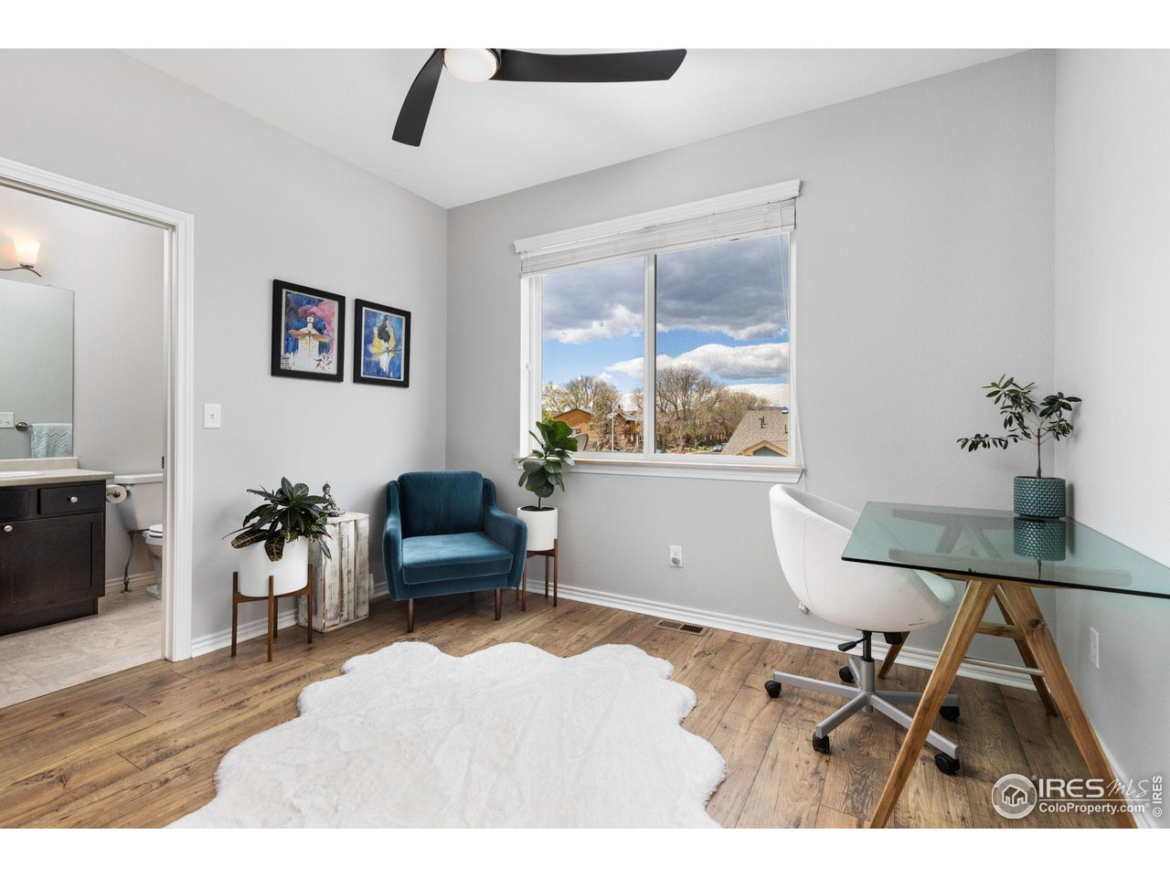 Furnished home office with a glass desk, teal armchair, and large window overlooking nearby rooftops.