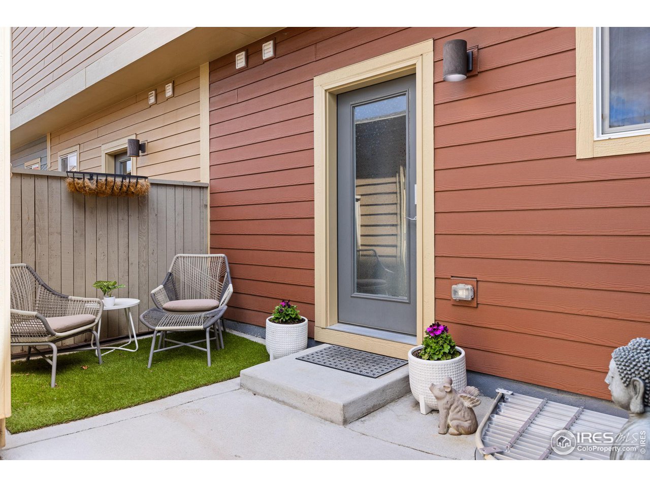 Home patio with woven chairs, artificial grass, and potted flowers beside a glass-paneled entry door.