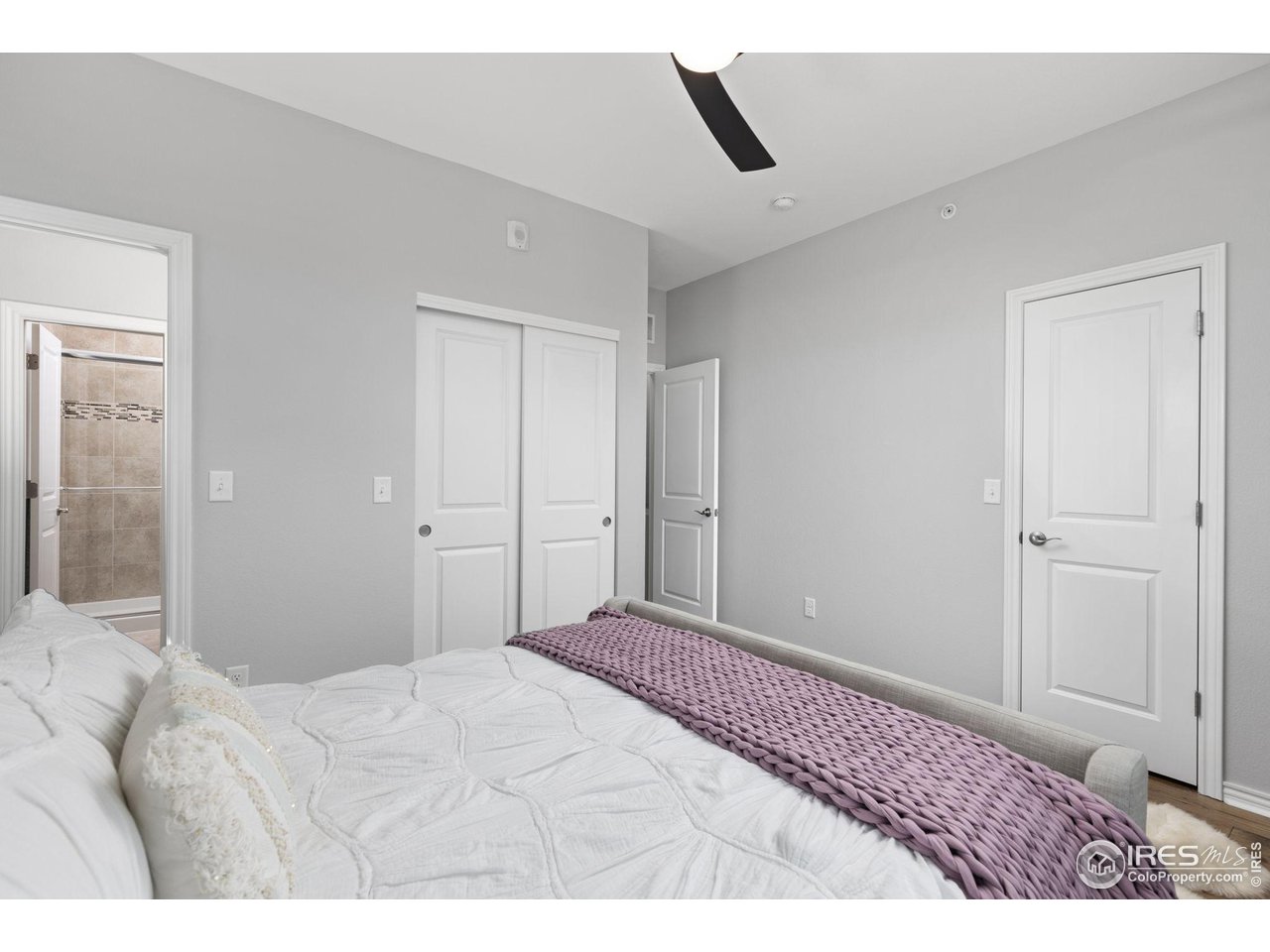 Bedroom with white bedding, a mauve knitted blanket, sliding closet doors, and an open view into a tiled bathroom.