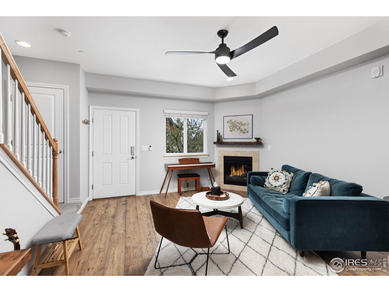 Living room with a teal sectional, lit fireplace, piano beneath the window, and staircase with wood handrail.