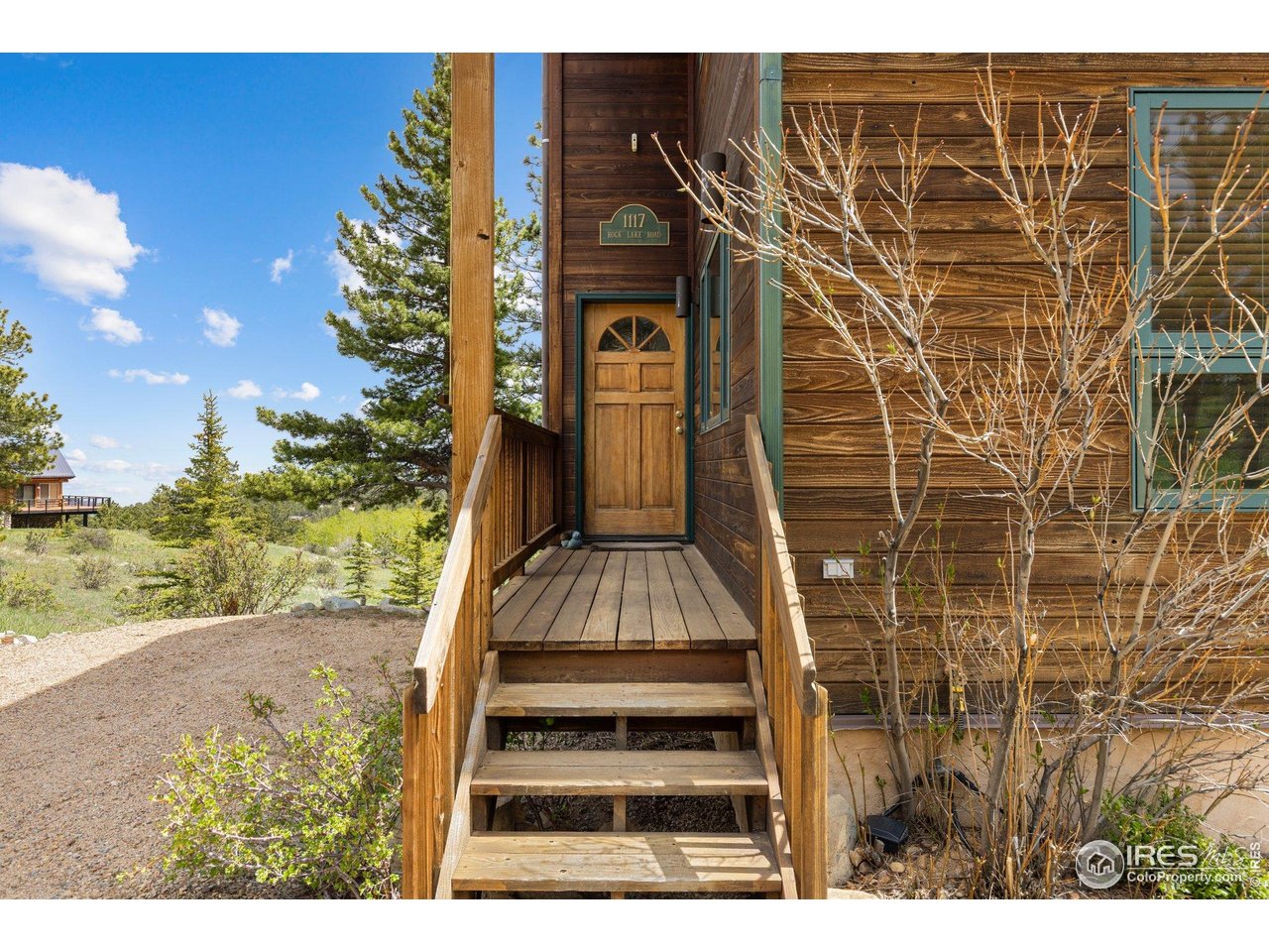 Wood-sided home entrance with a raised wooden porch, paneled door, and evergreens in the sunny landscape.