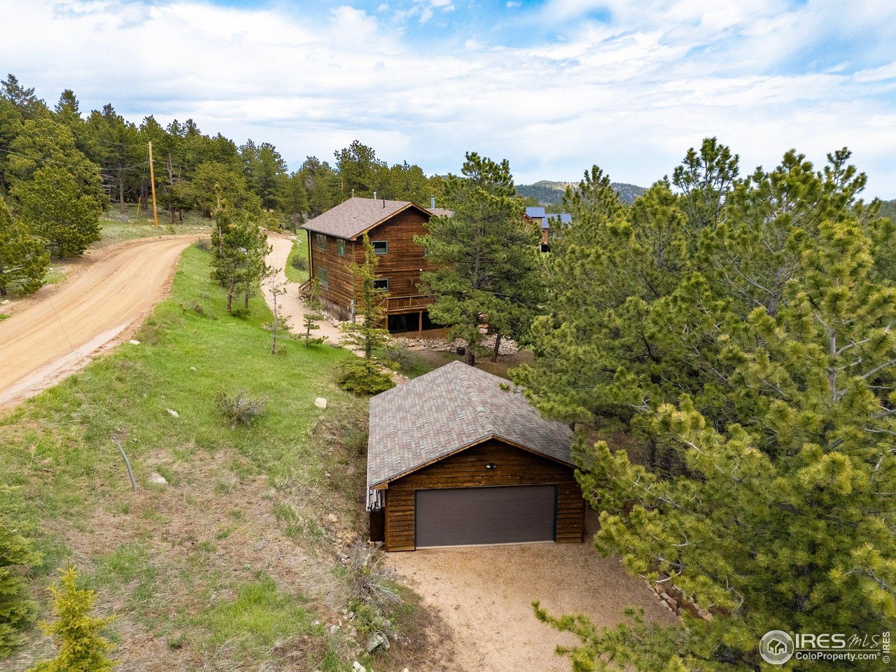 Aerial view of a wood-sided mountain home and detached garage surrounded by pine trees and a curving dirt road.