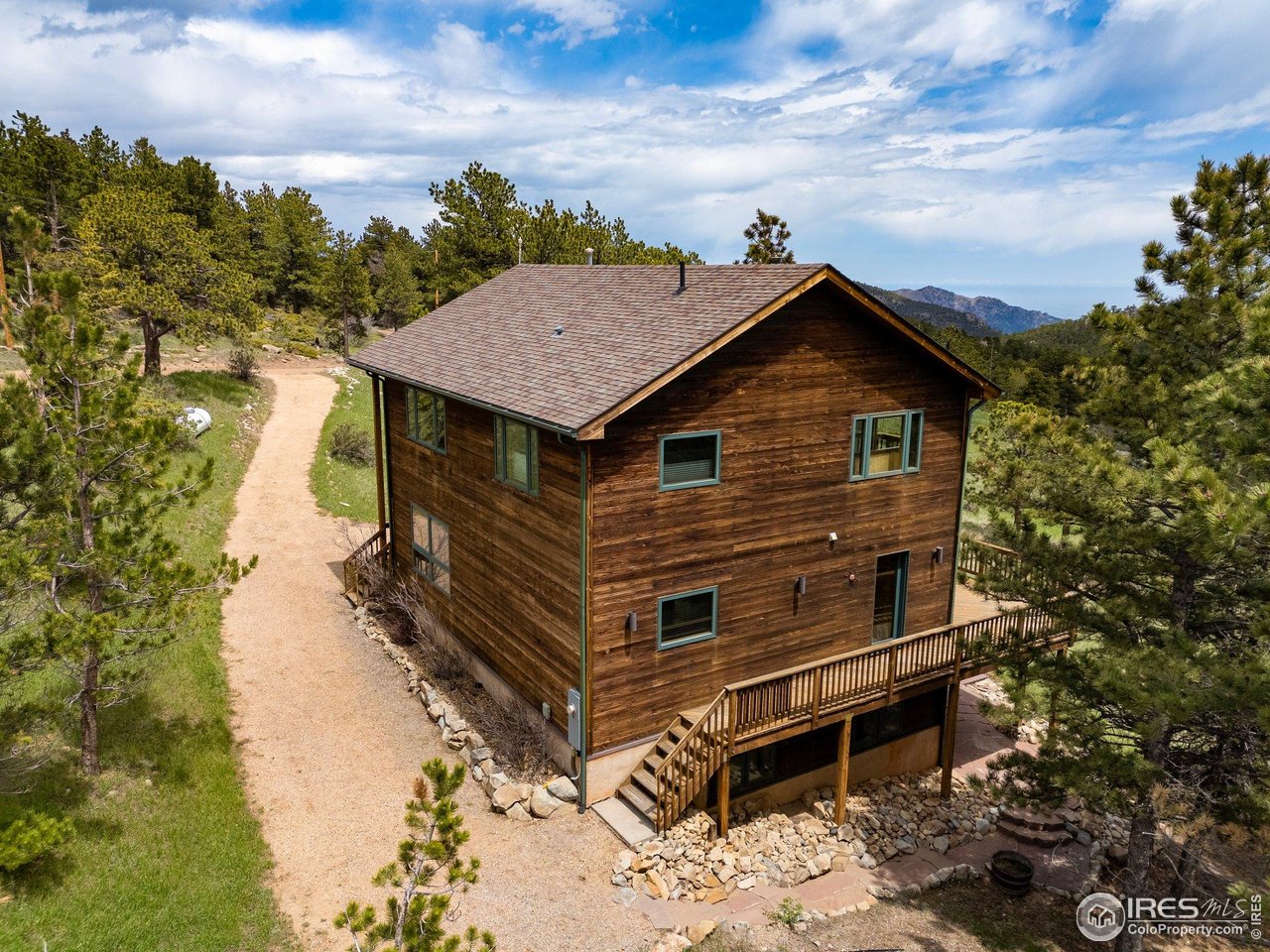 Wood-sided house with a raised deck among pine trees, gravel path, and distant mountains.