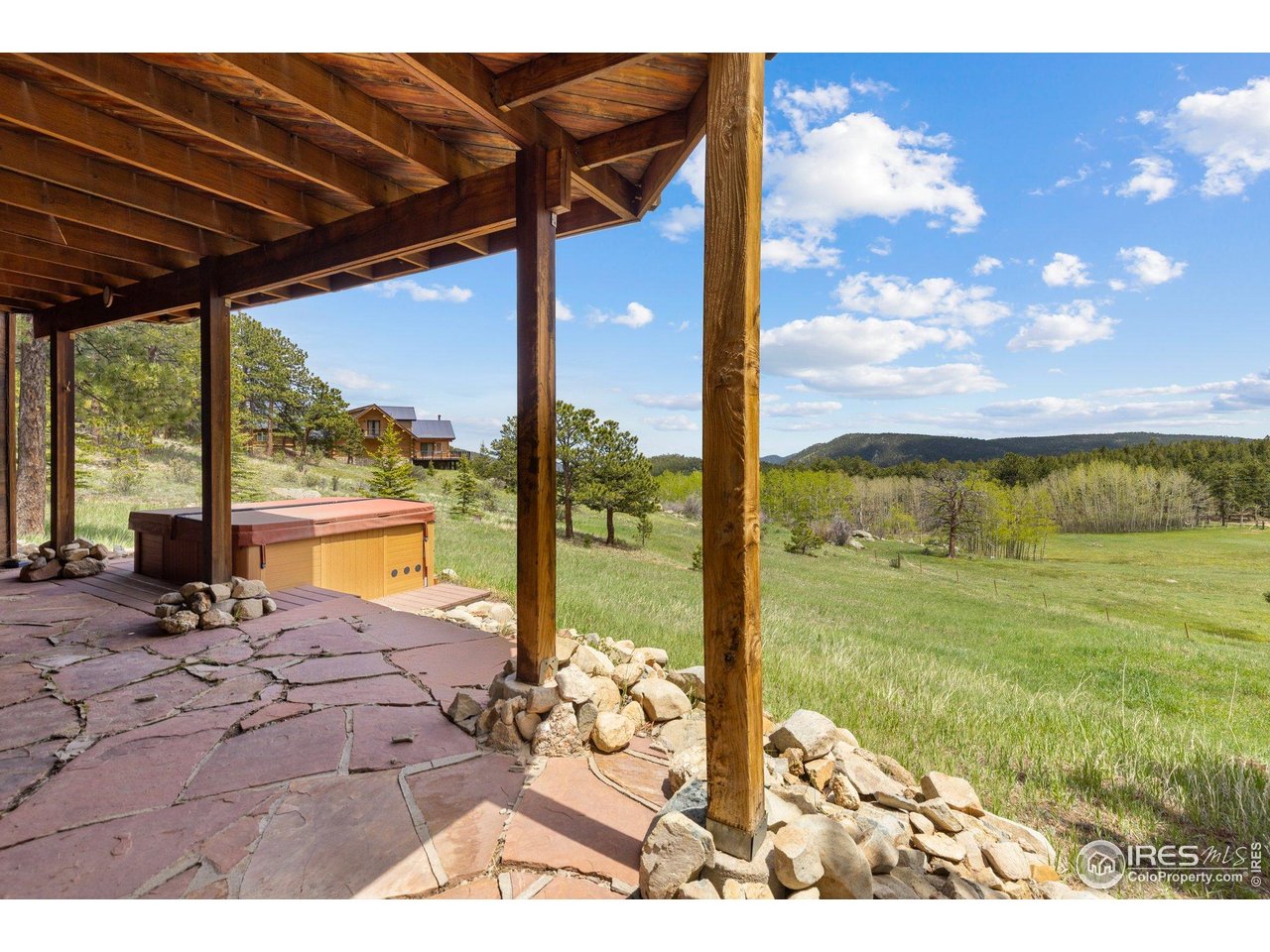 Covered flagstone patio and hot tub overlooking a grassy meadow and wooded mountain hills.