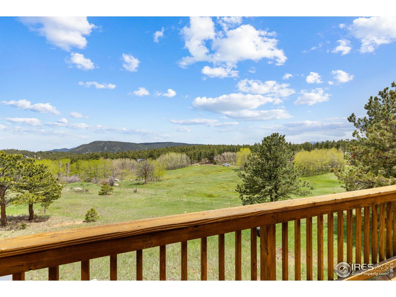 Meadow and forested hills viewed over a wooden deck railing beneath a blue, cloudy sky.