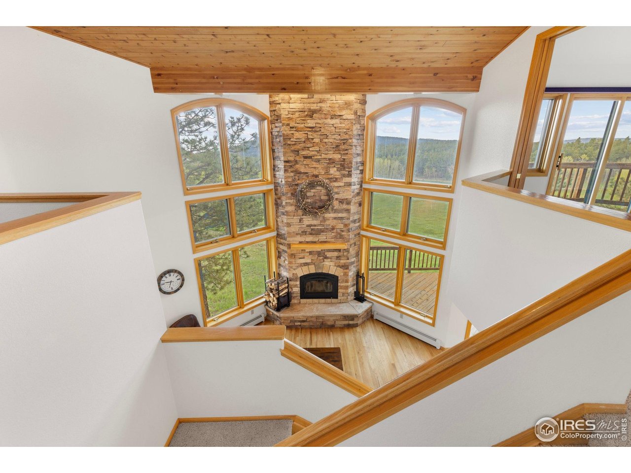 Tall living room with a stone fireplace, vaulted wood ceiling, and large windows overlooking wooded hills.