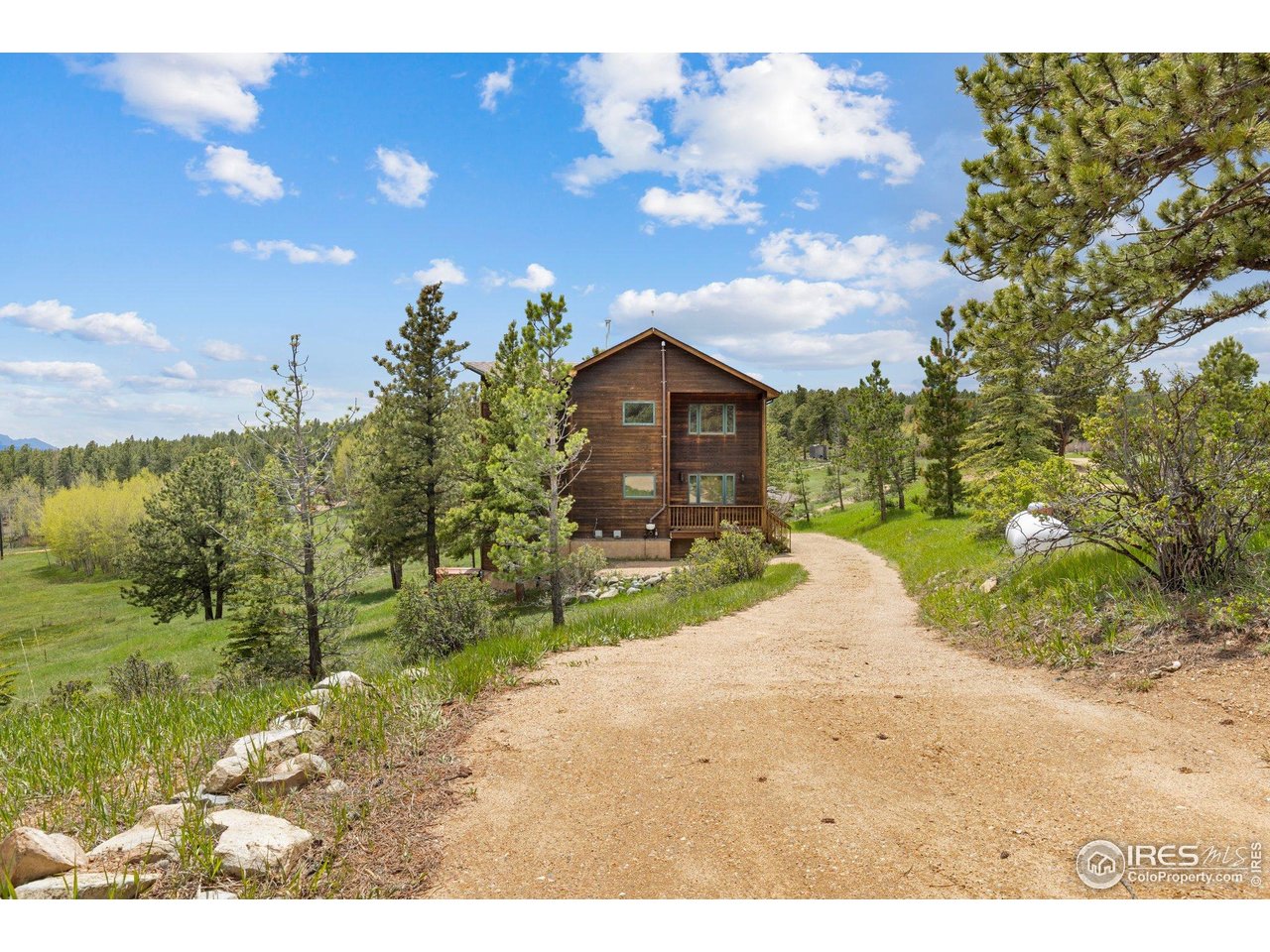 Wood-sided cabin among pine trees, with a gravel driveway and distant mountain ridge.