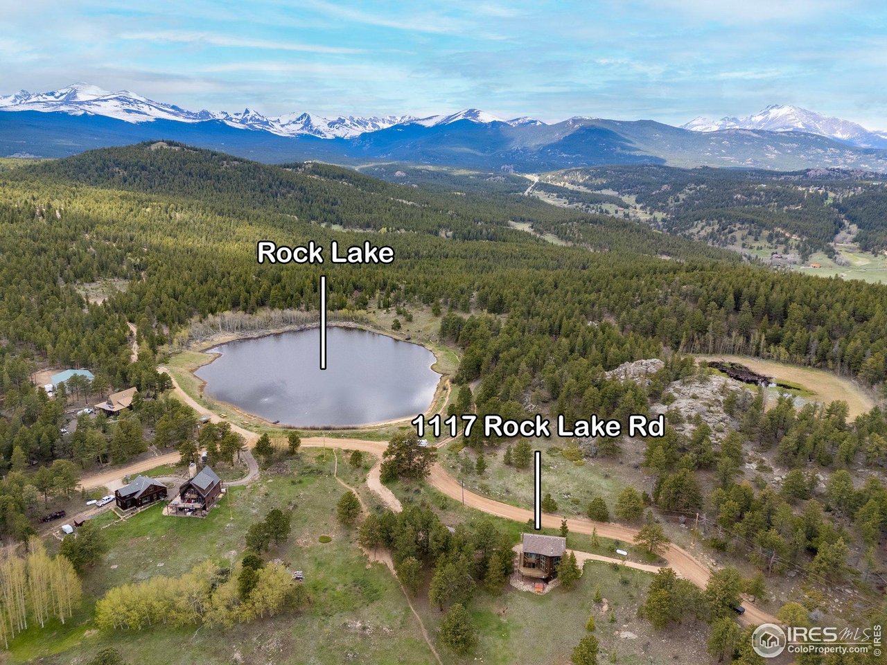 Aerial view of 1117 Rock Lake Rd beside Rock Lake, with forested hills and snow-capped mountains beyond.
