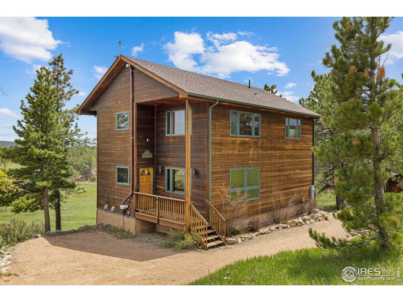 Two-story brown wood-sided home with a covered entry, porch steps, pine trees, and a gravel drive.
