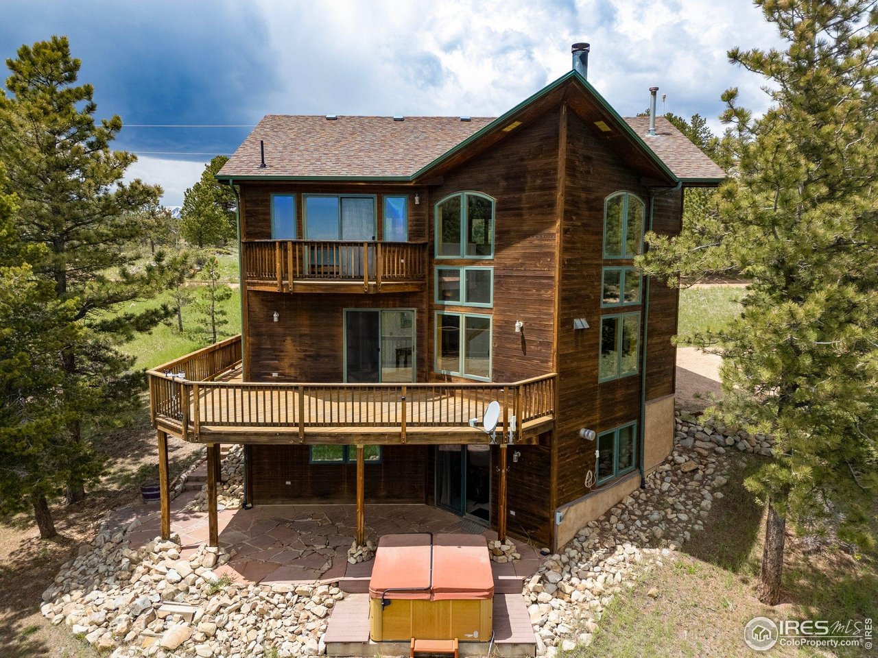 Two-story wood home with a wraparound deck and hot tub among pine trees.