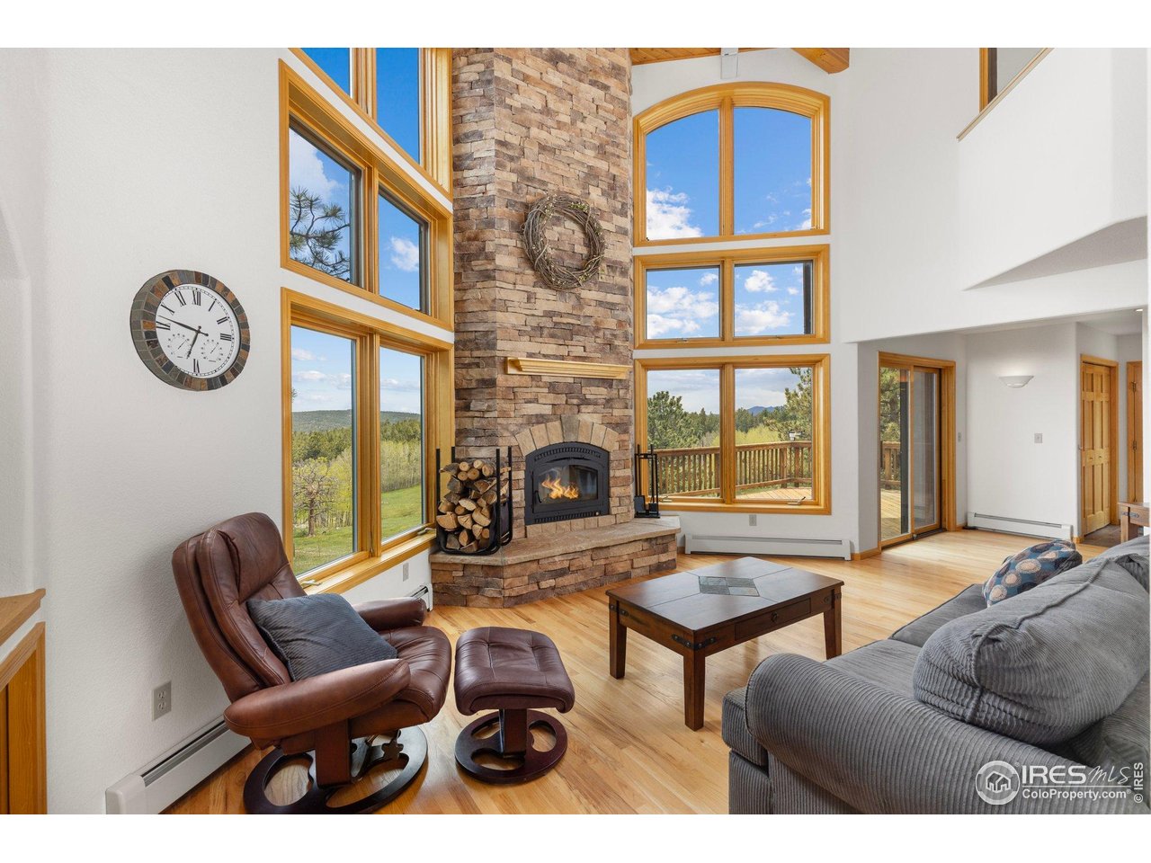Living room with a tall stone fireplace, vaulted ceiling, and large windows overlooking forested hills.