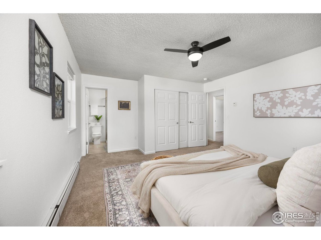 Furnished bedroom with a white bed, patterned rug, black ceiling fan, and open bathroom doorway.