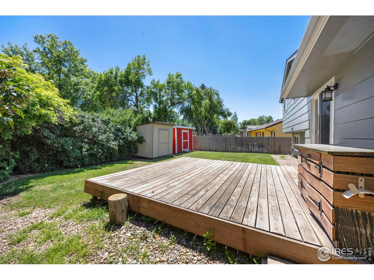 Backyard deck beside a hot tub, with lawn, leafy shrubs, and two storage sheds.