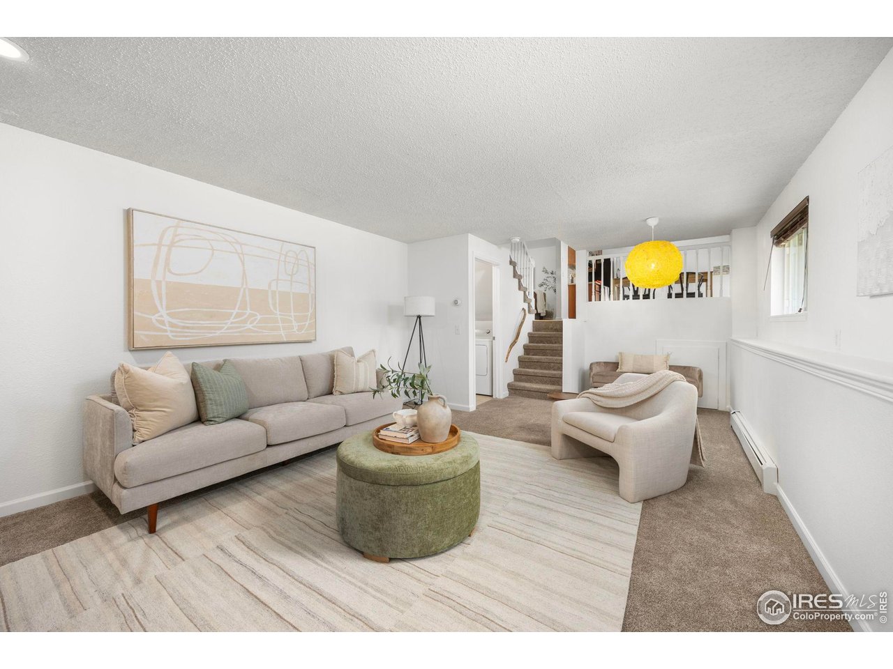 Staged living room with a beige sofa, green ottoman, pale rug, and staircase.