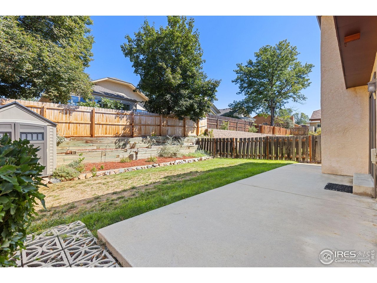 Backyard patio with a broad concrete slab, fenced lawn, tiered retaining walls, and mature trees.