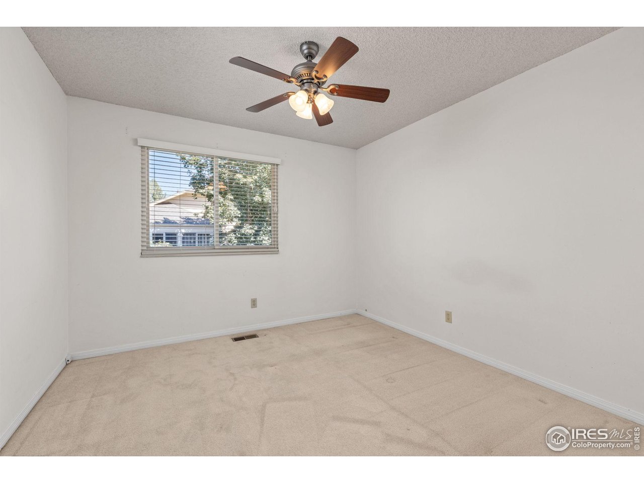Empty bedroom with beige carpet, window blinds, and a ceiling fan with lights.