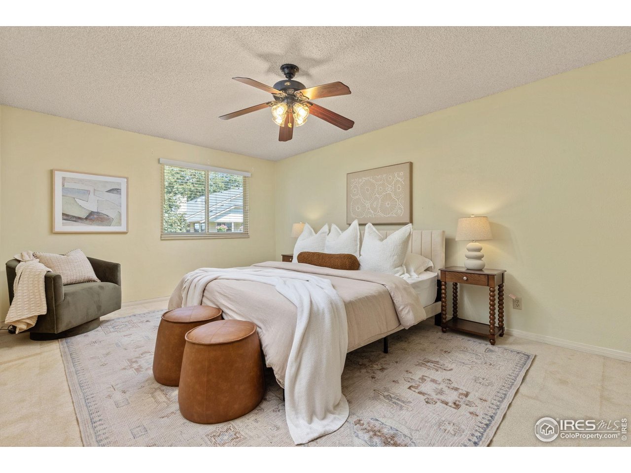 Staged bedroom with a white-and-beige bed, ceiling fan, window, upholstered chair, and brown ottomans.