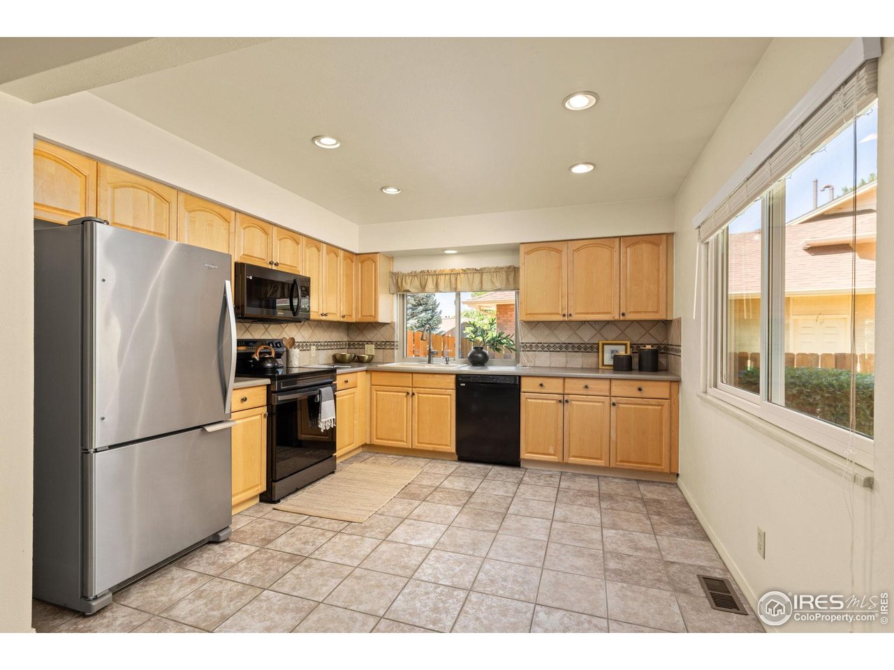 Kitchen with honey-colored cabinets, stainless-steel refrigerator, black appliances, and tiled floor.
