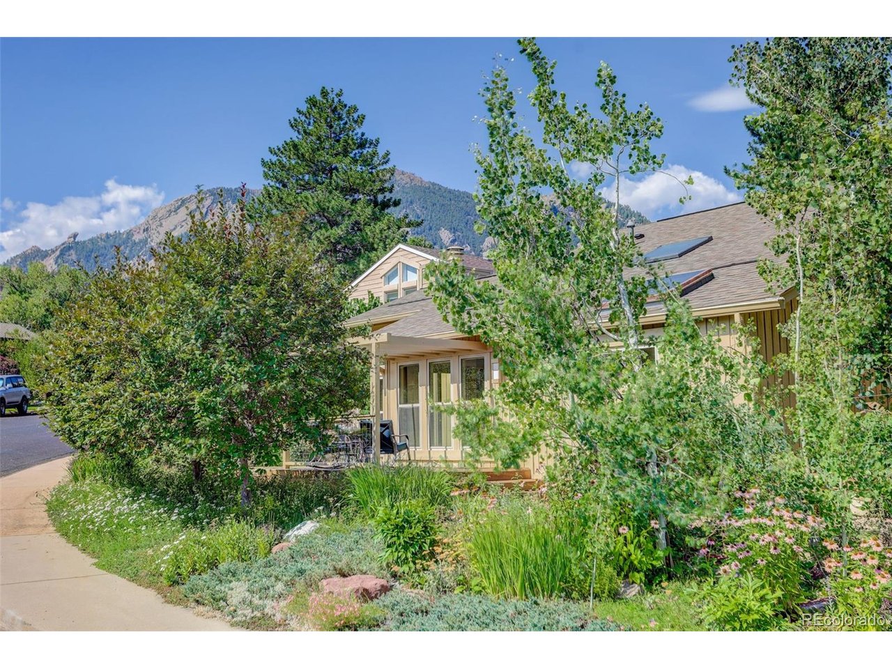 Tan multi-level house among leafy trees and flowers, with a mountain ridge behind it.