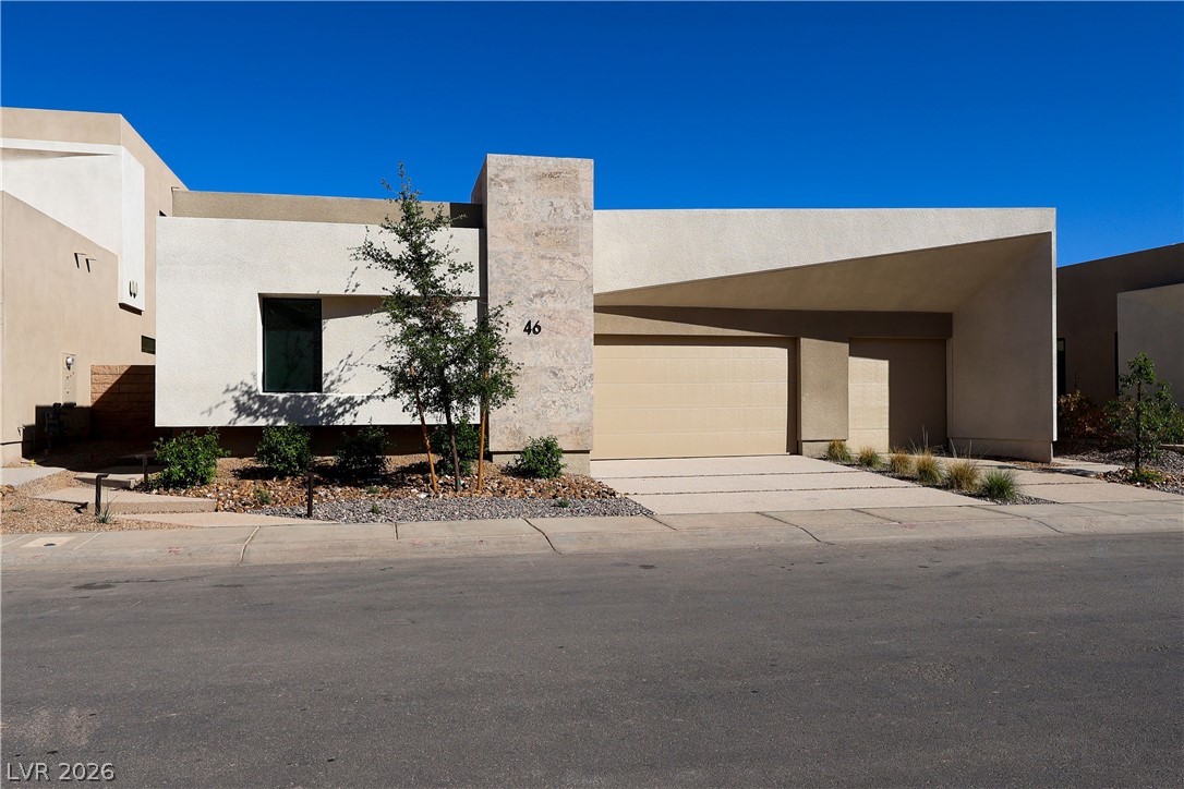Modern flat-roofed home with pale stucco, stone entry column, attached garage, and desert landscaping.