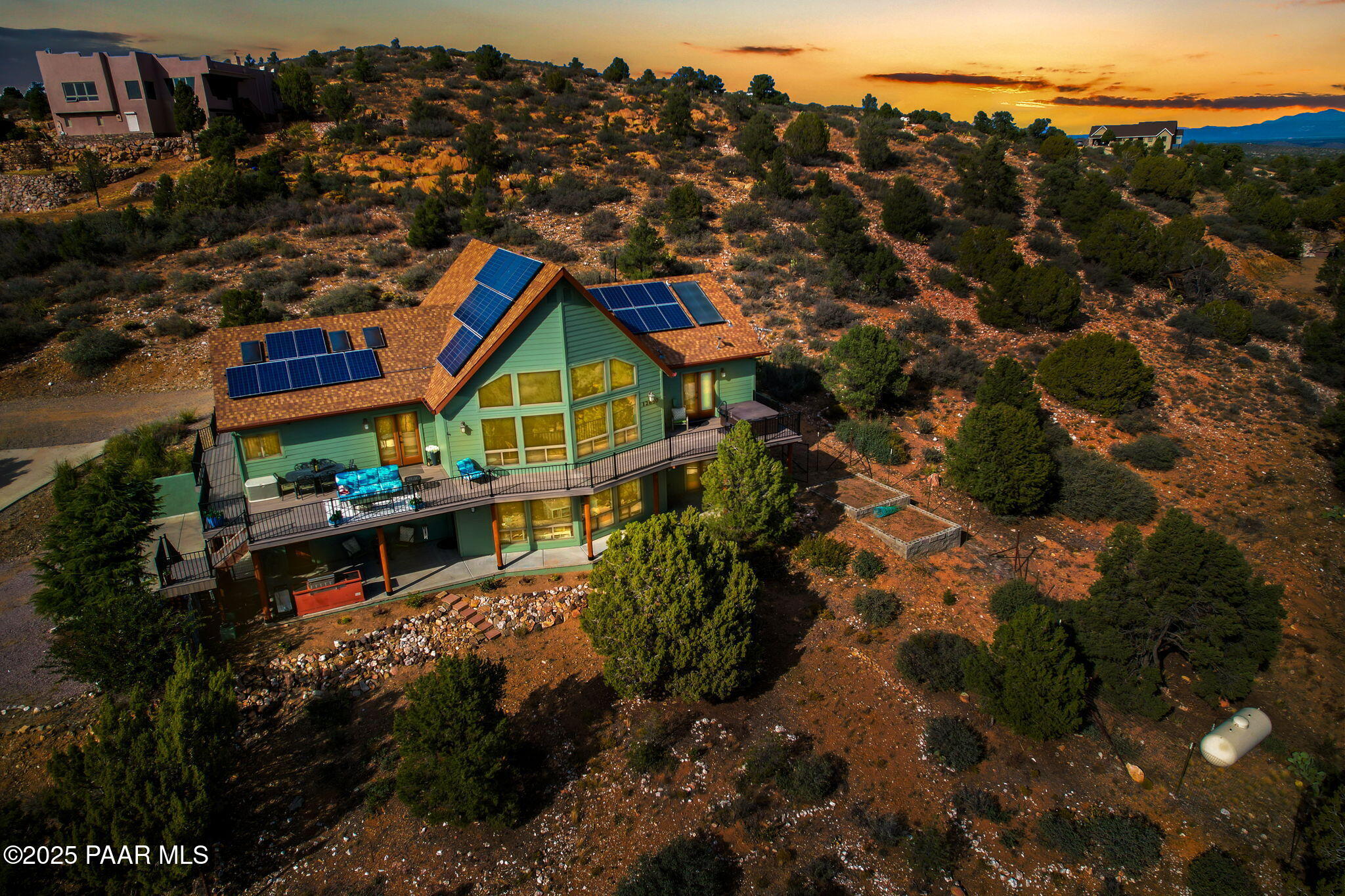 Hillside home with rooftop solar panels and broad decks amid desert scrub at sunset.