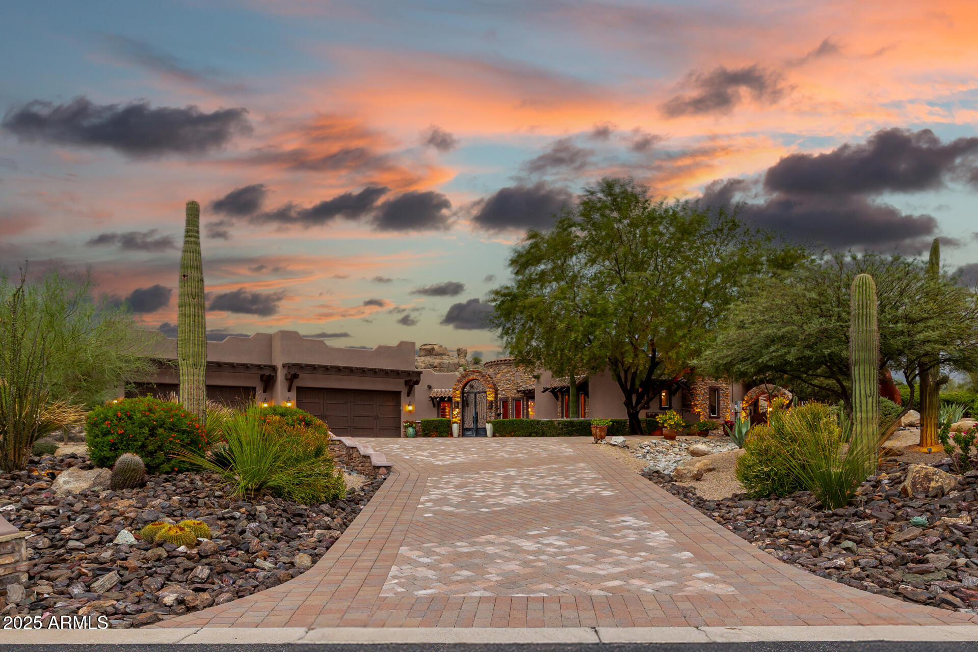 Southwestern-style home with stone accents and saguaro cacti along a broad paved driveway at dusk.