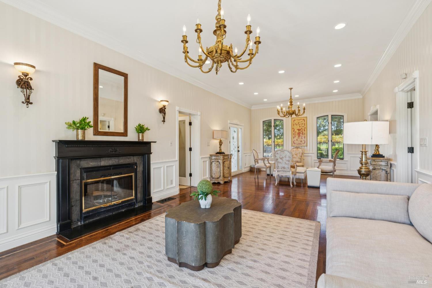 Living room with a black fireplace, brass chandeliers, and windows overlooking greenery.