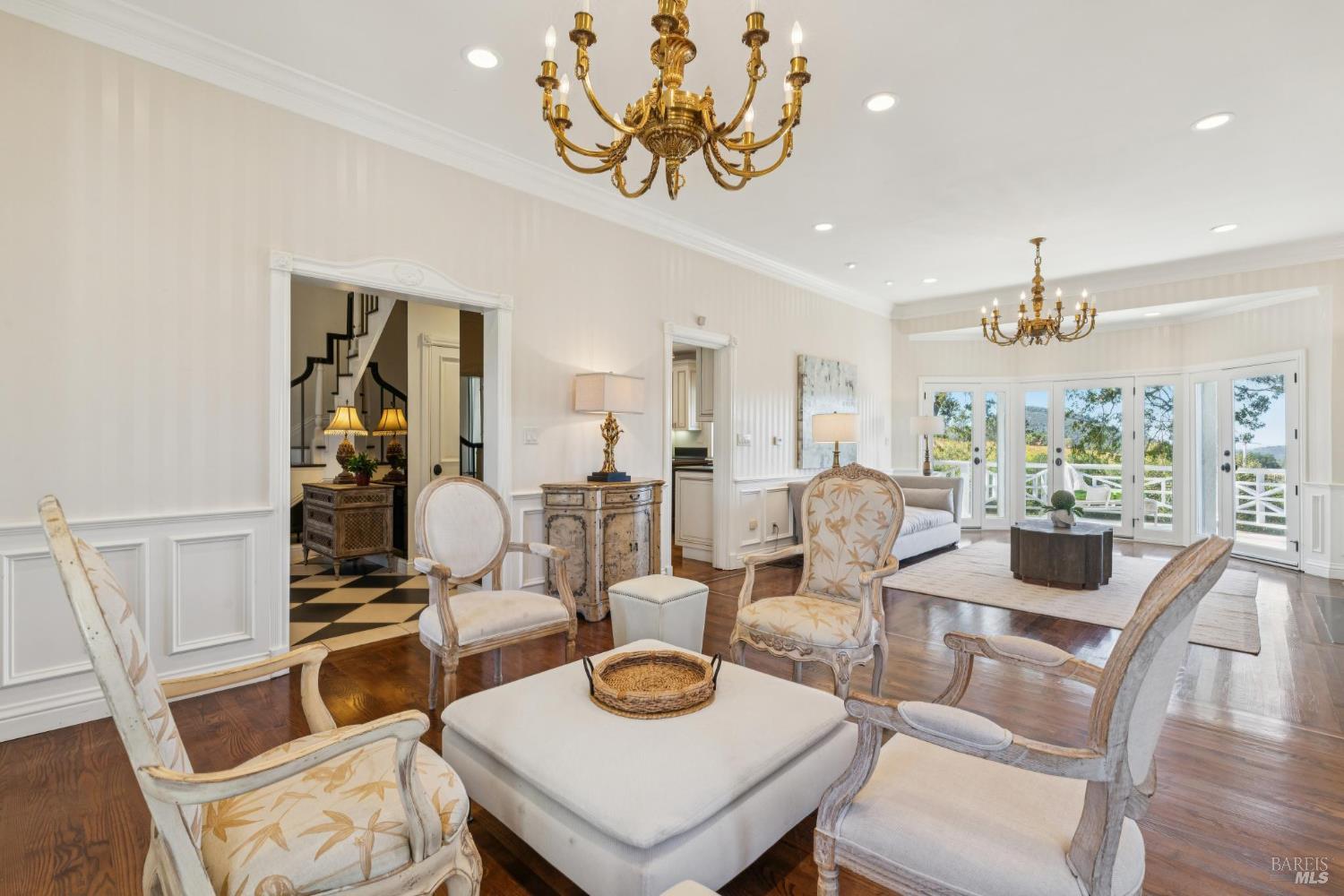 Living room with ornate chandeliers, upholstered seating, and French doors opening to a deck.