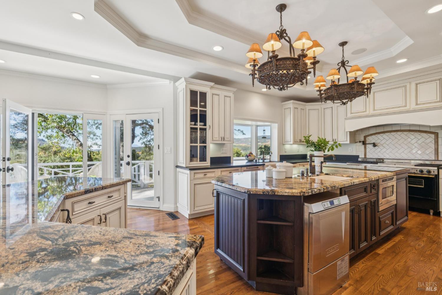 Kitchen with a dark wood island, granite counters, ornate chandeliers, and windows overlooking wooded hills.