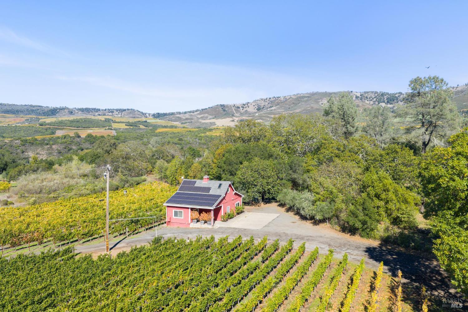 Aerial view of vineyard rows and a red solar-paneled outbuilding, with wooded hills in the distance.