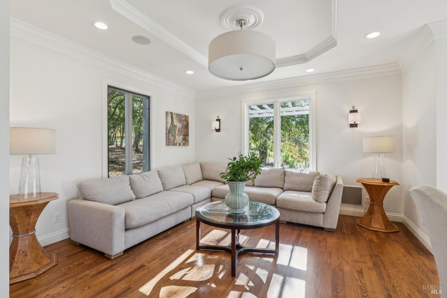 Living room with a light gray sectional, glass coffee table, hardwood floors, and windows overlooking trees.