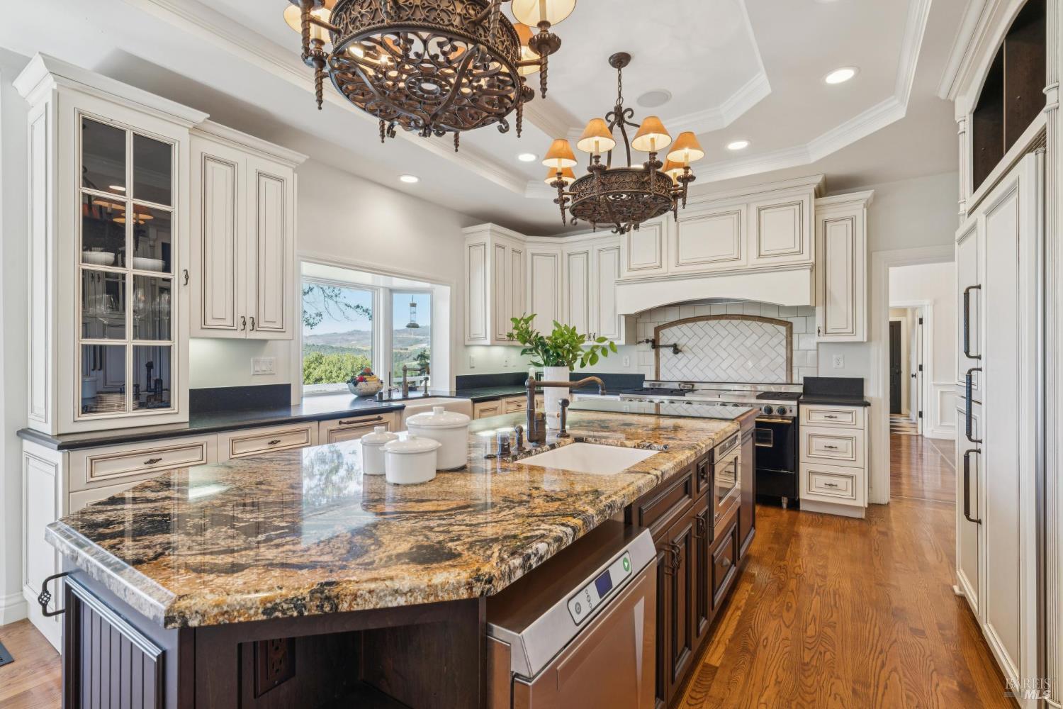 Kitchen with a large granite island, white cabinetry, ornate chandeliers, and wooded hills visible through the window.