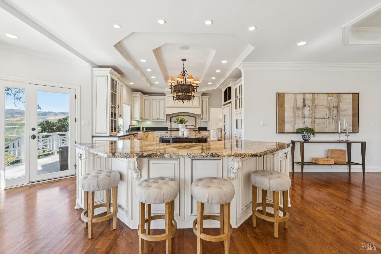Kitchen with a large granite island, four bar stools, white cabinetry, and vineyard views through glass doors.